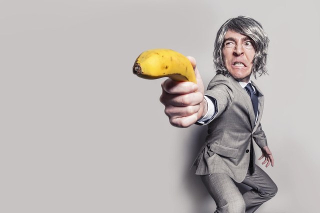 grey haired young man in grey suit defensively holding out a banana like a sword
